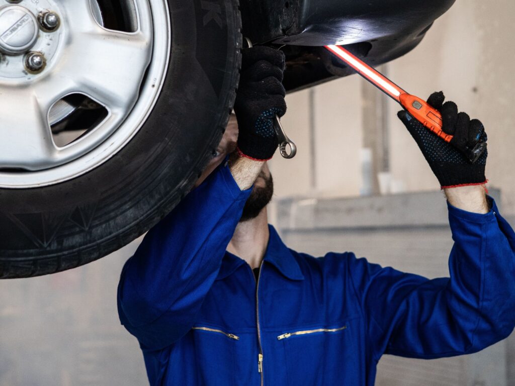 Service-1-min (1) Mechanic inspecting a car in an auto repair shop, checking under the vehicle with a flashlight.